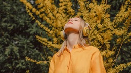 Das Beitragsbild des WDR3 Kulturfeature "Durch die Blume hören" zeigt eine Frau mit Kopfhörern vor einem gelb blühendem Baum