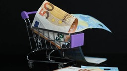 Das Beitragsbild des Dok 5 "Mag ich, kauf ich, gönn ich mir - Wirtschaftsfaktor Shopping" zeigt Geldscheine in einem kleinen Spielzeug Einkaufswagen auf schwarzem Hintergrund