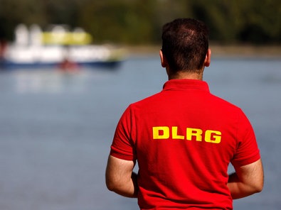 Ein Mitarbeiter der DLRG steht am Rheinufer, im Hintergrund ein Boot der Wasserschutzpolizei