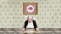 Ein Mann mit grauem Haar und Brille am Tisch, hält Messer und Gabel in den Händen, dahinter ein Foto mit Fleisch an der Wand.