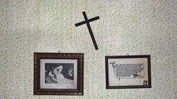 Ein schiefes Kreuz und zwei alte Bilder hängen an einer Wand.