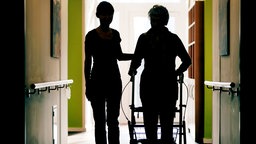 Eine Pflegekraft begleitet die Bewohnerin eines Altenheims mit Rollator durch den Flur.