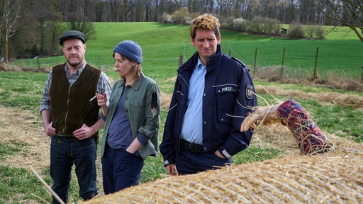 Ewald Schlichting (Roland Riebeling, l.), seine Frau Jutta (Milena Dreissig, 2.v.l.) und Heino Fuß (Sebastian Schwarz, r.) stehen auf dem Feld. Sie betrachten eine Leiche, die beim Heu einfahren in einen Strohballen geraten ist.
