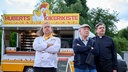 Hans Zielonka (Michael Hanemann, M.), Pastor Puttermann (Johannes Rotter, r.) und Hubert (Matthias Komm, l.) stehen vor Huberts Hähnchenwagen und schauen zum "Pascha Grill", wo die Hindus ihren Gottesdienst feiern.