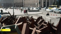 Im Vordergrund ein Haufen Metallkreuze im Hintergrund ein Platz in der Ukraine mit Fußgängern