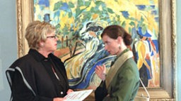 Dominique zeigt Helga das besagte Bild, dessen Preis bei der Auktion hochgetrieben werden soll.