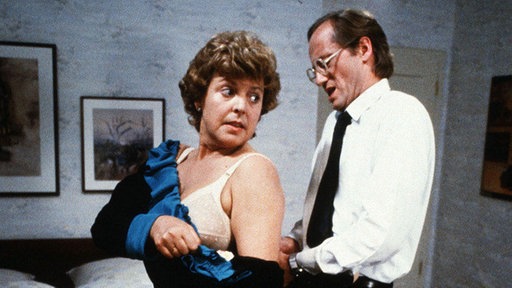 Hans (Joachim Hermann Luger) ist seiner Frau Helga (Marie-Luise Marjan) beim Ankleiden behilflich.