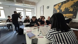 In einem Klassenzimmer mit einigen Schülerinnen und Schülern steht eine Person mit einer Kamera