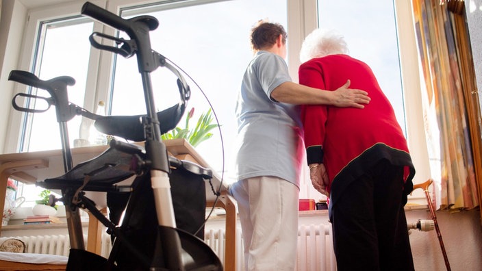 Eine Pflegerin und eine ältere Frau schauen zusammen aus einem Fenster, im Vordergrund ein Rollator
