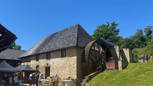 Vor einer Wassermühle stehen mehrere Menschen unter Zelten