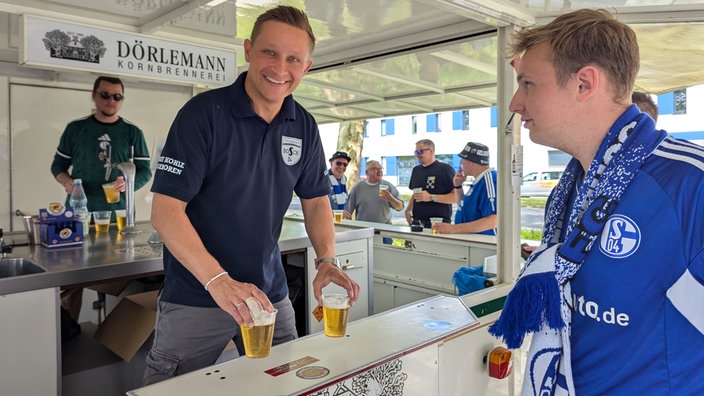 Schalkefan kauft zwei Bier an einem Bierstand. Bierverkäufer schaut lächelnd in die Kamera