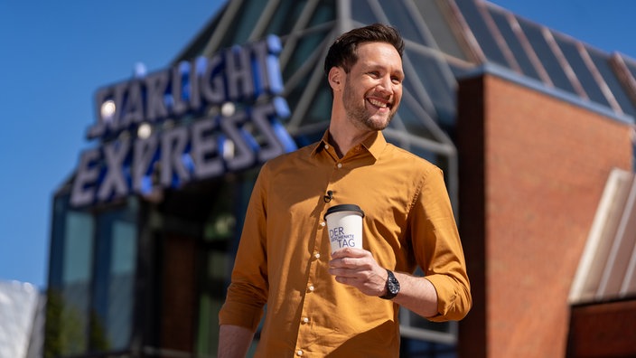 Moderator Sven Kroll hält einen Kaffebecher in der Hand und lächelt.