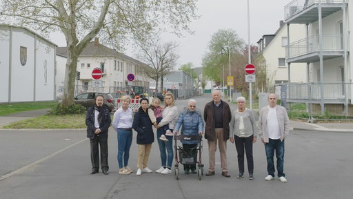 Neun Menschen verschiedenen Alters stehen auf einer Straße und schauen in die Kamera.