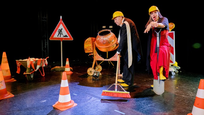 Zwei Opernsänger*innen in Arbeitskleidung mit Baustellen-Bühnenbild.
