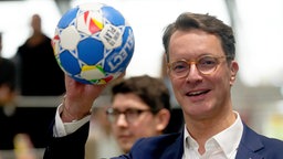 Ein Mann in Anzug und Brille mit einem blau-weißen Ball in der Hand.