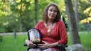 Frau in Rollstuhl sieht ernst in die Kamera. Sie hält eine bunte Urne in der Hand.