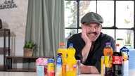 Doc Esser lacht in die Kamera. Um ihn herum befinden sich Getränke mit hohem Zuckergehalt.