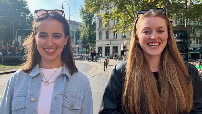 Zwei junge Frauen stehen nebeneinander auf einem städtischen Platz, beide lächeln in die Kamera. Im Hintergrund sind Bäume, Fahrräder und Gebäude zu sehen.