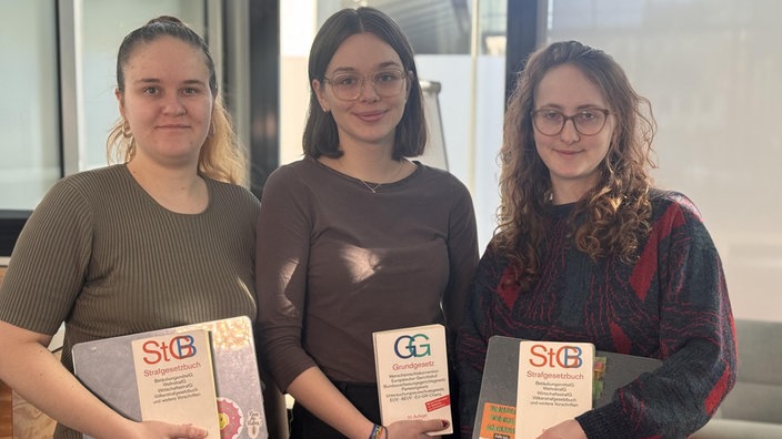 Porträtbild von Lilith Rein, Karla Steeb und Lilian van Rey. Sie lächeln in die Kamera und haben Strafgesetzbücher in der Hand. 