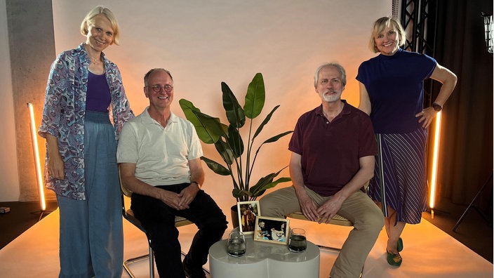 Zwei Frauen und zwei Männer stehen und sitzen gemeinsam in einem Studio vor warm beleuchtetem Hintergrund und blicken in die Kamera.