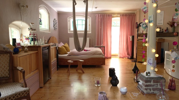 Heller Raum mit Holzfußboden, Bett, rosa Vorhängen und gemütlicher Dekoration. In der Mitte hängt ein Tragetuch von der Decke, Kerzen und Lampions sorgen für eine warme Stimmung.