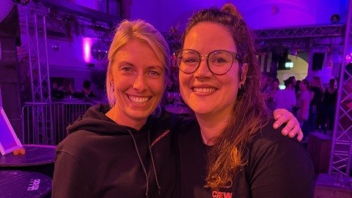 Zwei Frauen stehen Arm in Arm in einem Club bei bunter Beleuchtung und schauen lachend in die Kamera.