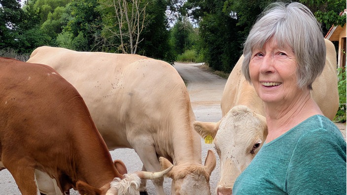 Ältere Frau mit grauen Haaren und grünem T-Shirt lächelt in die Kamera. Neben ihr stehen mehrere Kühe, die auf einer Landstraße fressen. Im Hintergrund Bäume und ländliche Umgebung. 