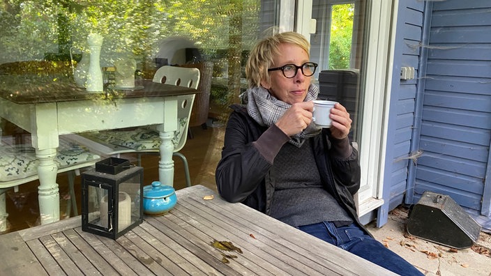Eine Frau mir kurzen blonden Haaren sitzt mit einer Tasse Kaffee an einem Holztisch auf der Terrasse ihres Hauses. 