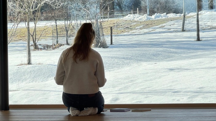 Eine Frau sitzt mit dem Rücken zur Kamera auf dem Boden vor einer großen Fensterfront und blickt nach draußen auf eine winterliche Landschaft.