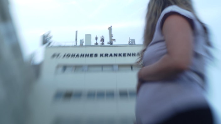 Unscharfes Foto einer schwangeren Frau im Vordergrund, die auf ein Krankenhausgebäude mit der Aufschrift "St. Johannes Krankenhaus"blickt.
