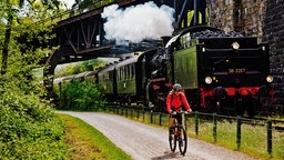 Eine Fahrradfahrerin fährt auf einem Radweg neben einer historischen Dampflok.