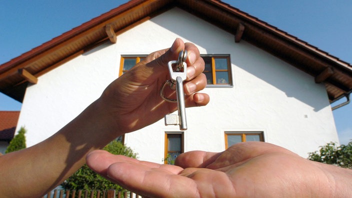 Vor einem Einfamilienhaus übergibt eine Hand einen Schlüssel in eine andere Hand