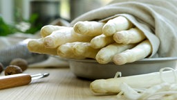 Weißer Spargel in ein Tuch gewickelt in einer Schüssel neben Sparschäler und einer geschälte Stange mit Schale daneben