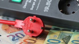 Ein roter Stecker vor einem Zeitschalter und Geldscheinen -  Symbolbild