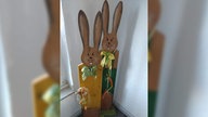 Zwei bemalte Osterhasen aus Holz stehen als Dekoration in einer Zimmerecke.