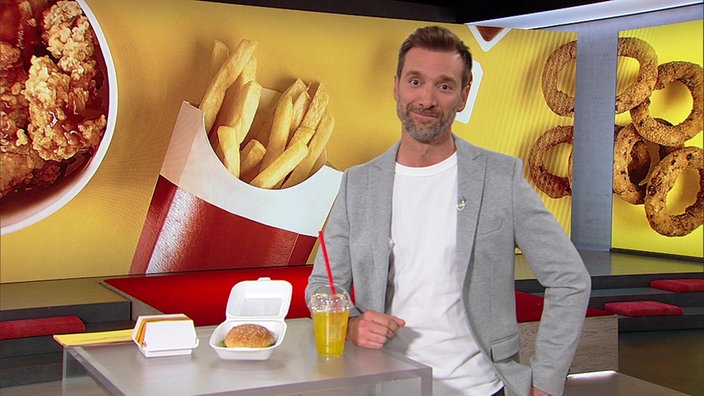 Daniel Aßmann steht im Markt-Studio. Sowohl neben als auch hinter ihm sind verschiedene Produkte der Fastfood-Industrie zu sehen.