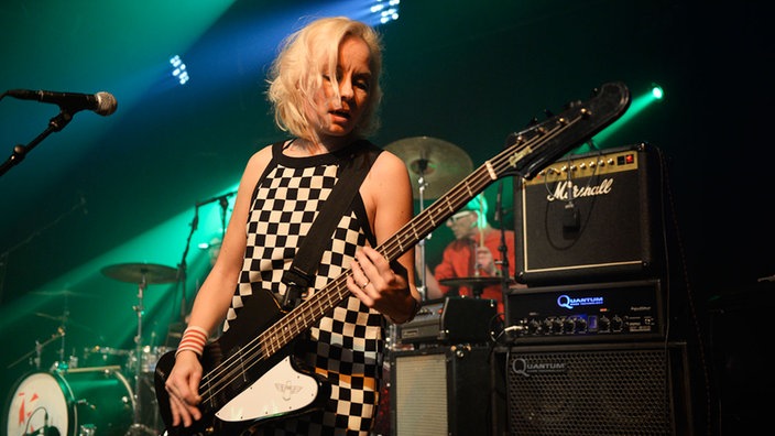Bassistin von Angelika Express mit ihrem Gibson Thunderbird E-Bass in schwarz mit weißem Schlagbrett, schaut verträumt aufs Griffbrett und spielt einen rockigen Groove mit einem Plektron