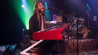 Keyboarder von Big Sugar steht hinter seinen zwei Keyboards, trägt eine lustige Brille und lange Dreads und singt ausgelassen ins Mikrofon