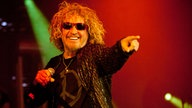 Der Sänger von Chickenfoot hat halb lange wallende Haareund trägt eine rote Sonnenbrille. In der rechten Hand hält er das Mikrofon mit dem linken Finger zeigt er ins Publikum