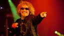 Der Sänger von Chickenfoot hat halb lange wallende Haareund trägt eine rote Sonnenbrille. In der rechten Hand hält er das Mikrofon mit dem linken Finger zeigt er ins Publikum