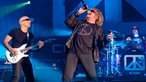 Sammy Hagar lehnt sich mit dem Mikrofon am Mund zurück links steht Joe Satriani an seiner Gitarre und im Hintergrund ist der Drummer an seinem Schlagzeug zu sehen