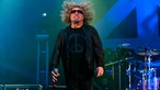 Sammy Hagar springt wodurch seine Haare hoch fliegen. In der rechten Hand hält er sein Mikrofon