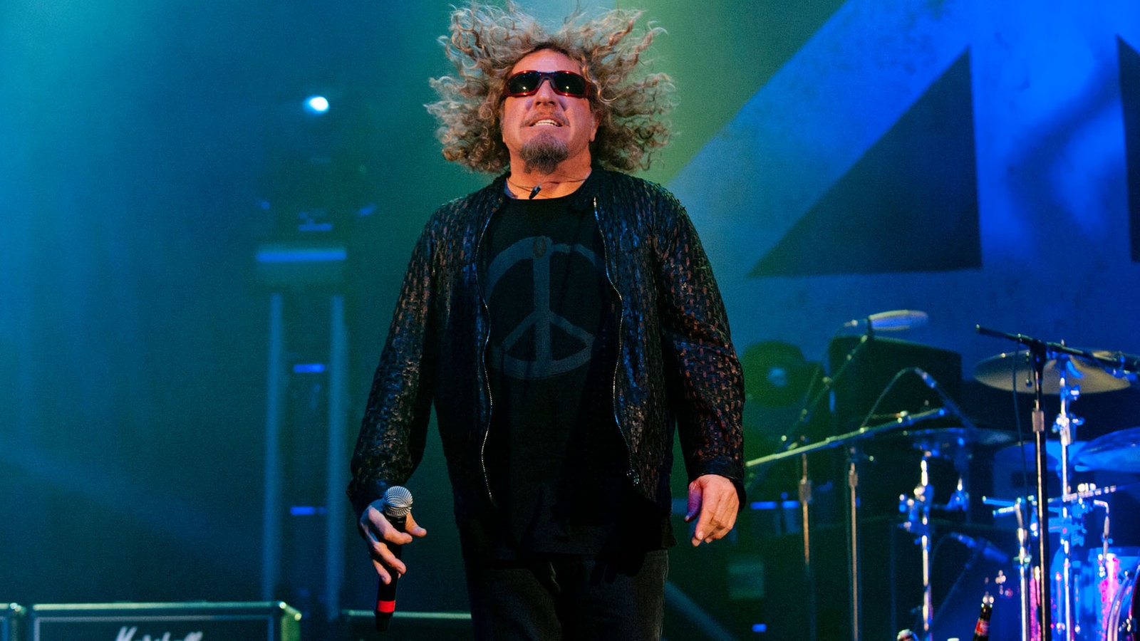 Sammy Hagar springt wodurch seine Haare hoch fliegen. In der rechten Hand hält er sein Mikrofon