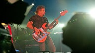 Michael Anthony am Bass im Vordergrund sieht man schwarze Silhouetten einer Person an einer Kamera