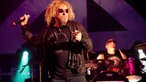 Sammy Hagar hat den rechten Arm mit dem Mikrofon in der Hand nach oben angewinkelt mit der linken Hand zeigt er in Richtung Publikum