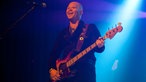 Bassist mit Glatze spielt und lacht im blauen Bühnenlicht.