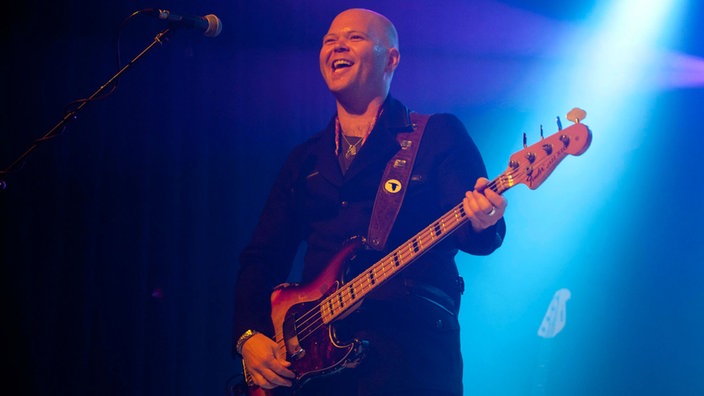 Bassist mit Glatze spielt und lacht im blauen Bühnenlicht.