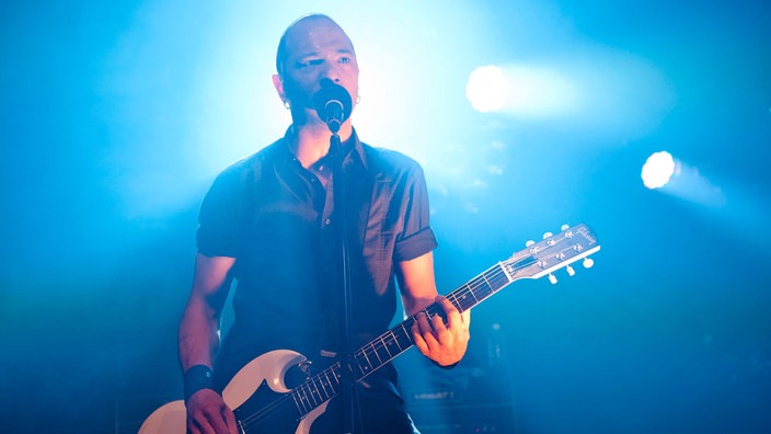 Danko Jones steht mit der Gitarre am Mikrofon und wird von hinten blau angestrahlt.