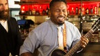 Mann mit verbissenem Gesicht beim Gitarre spielen, hinter ihm steht ein Mann mit Vollbart