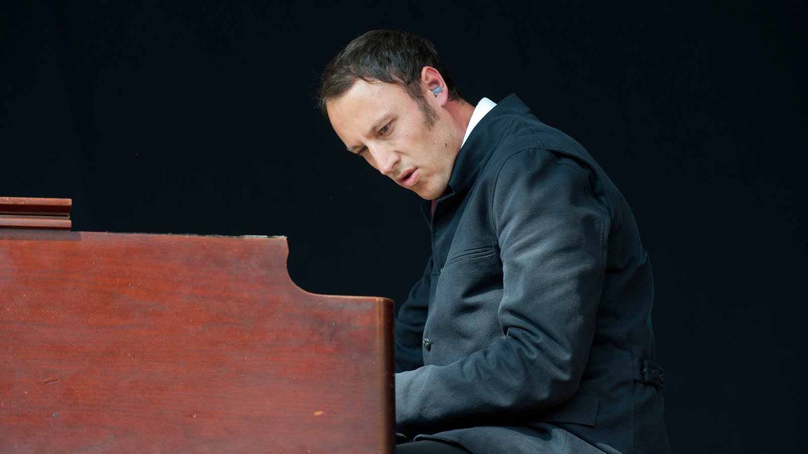 Mann im schwarzen Mantel sitzt an einer Orgel und schaut nach unten